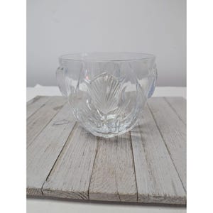 May include: A clear, decorative glass bowl with a leaf design. The bowl has a rounded shape with a wide opening and two small handles. The leaf pattern is etched into the glass, creating a textured surface. The bowl is sitting on a wooden surface.