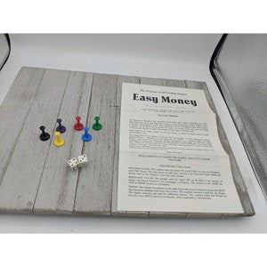 Op de afbeelding: Een vintage bordspel genaamd "Easy Money" met een speelbord, dobbelstenen en speelstukken. Het speelbord is gemaakt van hout en heeft een lichtbruine kleur. De speelstukken zijn kleurrijk en omvatten rood, blauw, groen, geel en paars. De dobbelstenen zijn wit met zwarte stippen. De spelinstructies zijn afgedrukt op een witte kaart en bevatten de tekst "Easy Money" en "The Greatest of All Trading Games!"