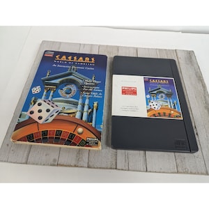 May include: A black and white game cartridge for the Philips Interactive World of Gambling Caesars World of Gambling game. The cartridge has a picture of a casino building with a roulette wheel and dice on the front. The text "Caesars World of Gambling" is printed on the front of the cartridge.