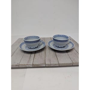 May include: Two matching teacups and saucers with a blue and white floral pattern. The teacups have a handle and sit on round saucers. The design includes a geometric border and small dot details. The set is on a light-coloured wooden surface.