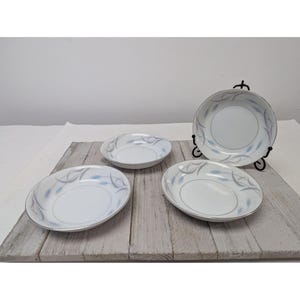 May include: A set of four white ceramic bowls with a silver rim and a delicate blue and gray floral design. One bowl is displayed on a black metal stand. The bowls are arranged on a weathered wooden surface.