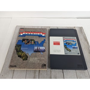 May include: A black and gray Rand McNally's America United States Atlas book with a map of the United States divided into sections for people, environment, and economy. The book is next to a black Philips Interactive Atlas of America CD-ROM case.