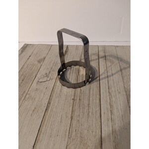 May include: A silver-colored metal biscuit cutter featuring a circular base with a scalloped edge and a tall, arched handle attached to the sides. The tool is constructed from a single strip of metal bent into a loop with a handle extending upward.