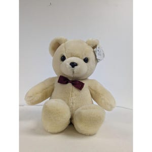 May include: A cream-coloured plush teddy bear wearing a burgundy bow tie. The bear is sitting with its paws in front of it.