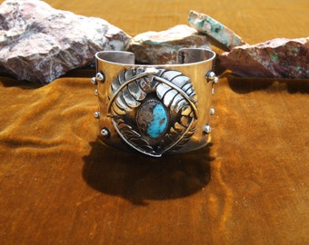 May include: A silver cuff bracelet with a turquoise stone set in a leaf design. The bracelet is decorated with small silver beads.