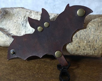 May include: A dark brown leather bat-shaped keychain with brass rivets and a black clasp. The bat has pointed ears and detailed wing edges. The keychain is designed to attach to keys or a bag.