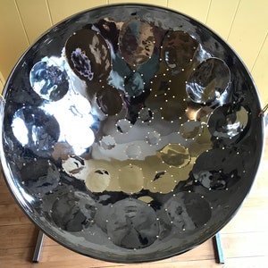 May include: A silver steel pan drum with a circular shape and a shiny surface. The drum has a series of round indentations on the surface, each with a small metal dot in the center. The drum is mounted on a stand with two legs.