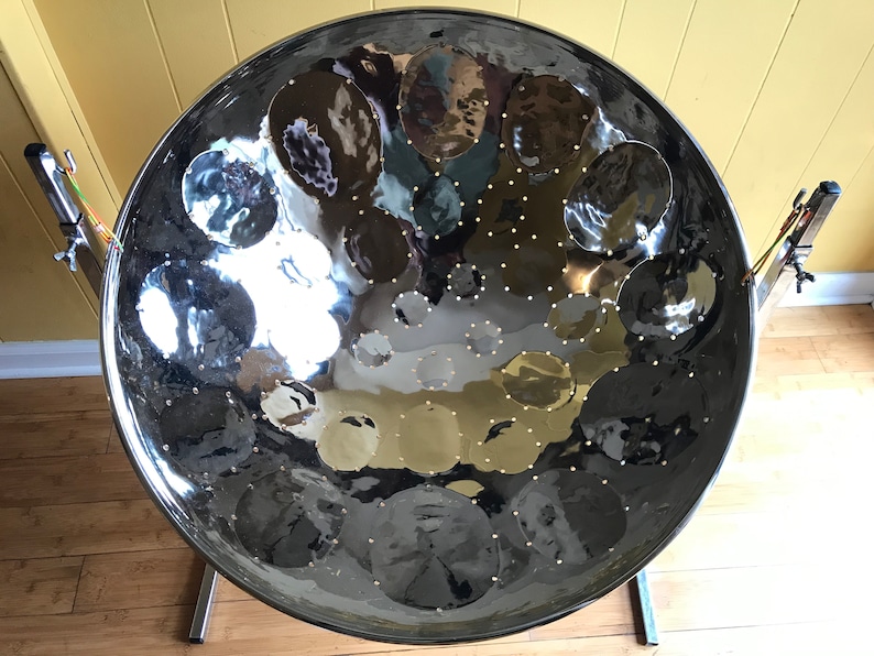 May include: A silver steel pan drum with a circular shape and a shiny surface. The drum has a series of round indentations on the surface, each with a small metal dot in the center. The drum is mounted on a stand with two legs.