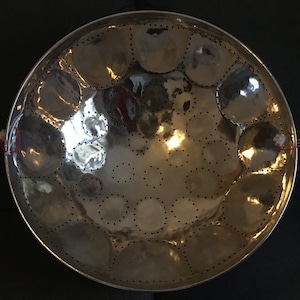 May include: A silver steel pan with a circular shape and multiple round indentations. The pan has a shiny, reflective surface and is decorated with small holes around the edges of the indentations.
