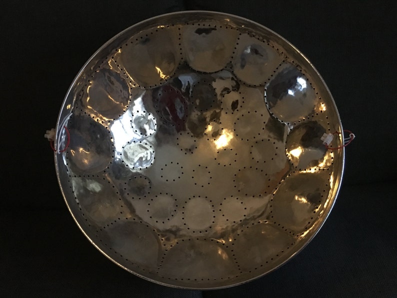 May include: A silver steel pan with a circular shape and multiple round indentations. The pan has a shiny, reflective surface and is decorated with small holes around the edges of the indentations.