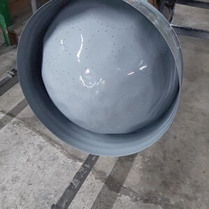 May include: A gray metal colander with small holes hanging from a hook. The colander is suspended above a concrete floor.