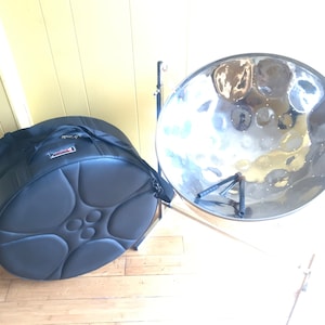 May include: A black padded case for a steel pan drum, with a steel pan drum on a stand in the background.
