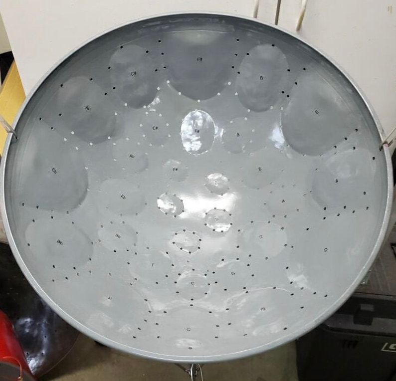 May include: A steel pan drum with a gray finish and a variety of notes labeled on the surface. The notes include C, D, E, F, G, A, and B.