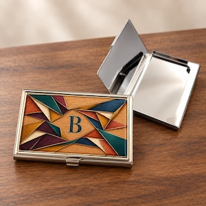 May include: A rectangular metal business card holder with a hinged lid. The front features a geometric inlay of wood and colored segments in teal, orange, and purple, centered with the letter B. One holder is closed, and one is open.