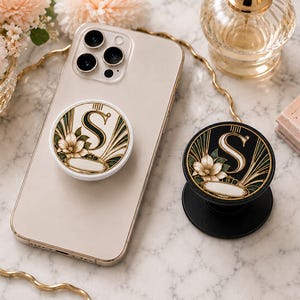 May include: Two circular phone grips featuring a decorative letter S, floral accents, and gold-toned geometric lines. One grip has a white base attached to a smartphone, while the other has a black base. Both display a matching botanical design.