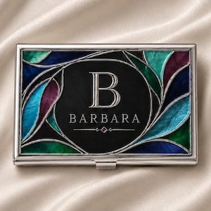 May include: A rectangular silver-toned metal business card case featuring a central black oval panel with the letter B and the name BARBARA engraved in silver. The surrounding border displays a stained-glass pattern with blue, teal, and purple.