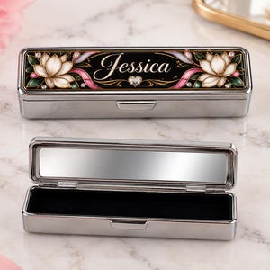 May include: A rectangular silver metal case with a hinged lid featuring a black background, white lotus flowers, pink ribbons, and the name Jessica in white script. The interior contains a rectangular mirror and a black velvet-lined base.