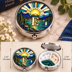 May include: A round silver metal pill case with a stained glass style lid featuring a sun, mountains, and the letter E. The case measures 1.75 inches in diameter and has three internal compartments containing white, tan, and green pills.
