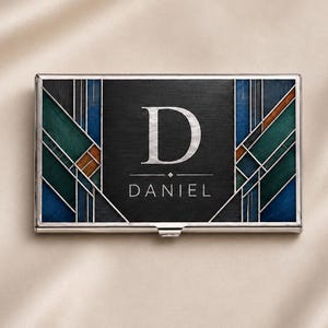 May include: A rectangular metal business card holder featuring a black center panel with the letter D and the name DANIEL engraved in silver. The sides display a geometric pattern of blue, teal, and brown segments separated by thin silver lines.