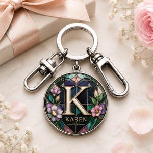 May include: A silver-toned metal keychain featuring a circular pendant with a stained-glass style design. The pendant displays a large letter K, the name KAREN, and floral motifs in shades of purple, pink, and green against a dark background.