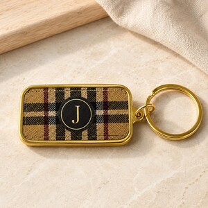 May include: A rectangular keychain featuring a tan, black, and red plaid pattern. In the center, a circular black emblem displays the gold letter J. The keychain is encased in a polished gold-tone metal frame and includes a matching gold-tone ring.