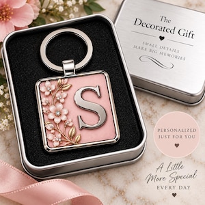 May include: A square metal keychain featuring the letter S in silver, accented with pink and gold floral details. The keychain is presented in a silver-colored metal gift box with the text The Decorated Gift, Small details make big memories.