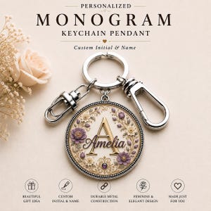 May include: A silver-toned circular keychain pendant featuring a gold-colored letter A and the name Amelia in script. The pendant is decorated with purple and cream-colored floral motifs and a small purple gem. It includes a metal ring and two clasps.