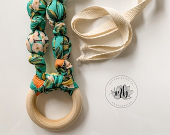 May include: A teal and orange fabric baby teether with a wooden ring and a white cotton strap. The teether has a floral pattern and the strap has a "Mother Bliss Co. Handcrafted" logo.
