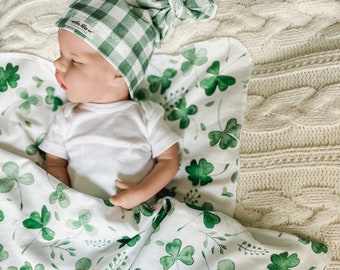 May include: A white baby blanket with a green shamrock pattern. The blanket is made of soft, breathable fabric and is perfect for cuddling. The blanket is handmade and features the text "Mother Bliss Co. Handcrafted". A baby is wearing a green and white gingham knot hat.