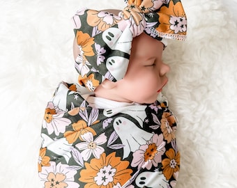 May include: A baby wearing a swaddle with a floral and ghost print. The swaddle is in shades of green, orange, pink, and white. The baby is wearing a matching headband with a large bow.