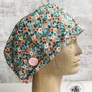 May include: A blue scrub hat with a floral print featuring pink and white flowers, green leaves, and gold accents. The hat has a pink button and a tie.