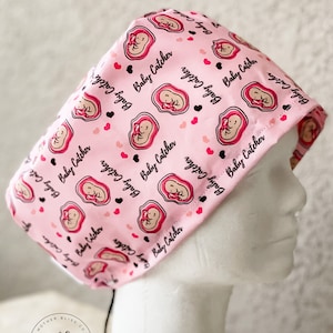 May include: Pink surgical scrub hat with a repeating pattern of a stylized pregnant woman and the text "Baby Catcher". The hat has a black drawstring and a small logo that says "Mother Bliss Co. Handcrafted".