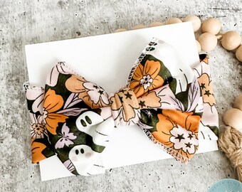 May include: A large, orange, pink, and green floral print bow with white ghost silhouettes. The bow is tied with a knot in the center and has a white background.