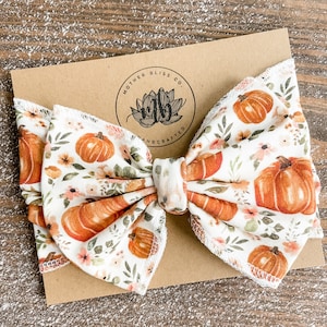 May include: A white fabric bow with a pumpkin and floral print. The bow is tied with a white ribbon and has a brown tag with the text "Mother Bliss Co. Handcrafted".