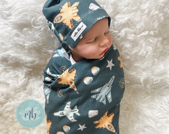 May include: A baby swaddled in a dark green swaddle with a white star and airplane print. The baby is wearing a dark green and yellow airplane print knotted beanie with the text "Mother Bliss Co." on the side.