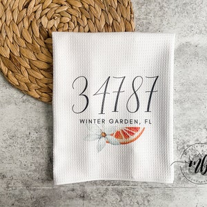 May include: White waffle-weave kitchen towel with the numbers "34787" and the text "WINTER GARDEN, FL" in black script. A watercolor illustration of a citrus fruit and flower is below the text. A woven placemat is in the background.