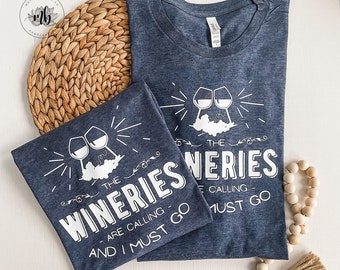 May include: Two navy blue t-shirts with a white graphic design of wine glasses and the text "The Wineries Are Calling And I Must Go".