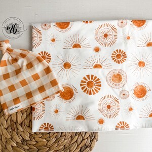 Puede incluir: Un gorro de beb&eacute; de cuadros blancos y naranjas con un nudo en la parte superior y una manta de arrullo blanca con un patr&oacute;n repetido de soles naranjas. El texto "maple bliss" est&aacute; en la etiqueta de la manta.