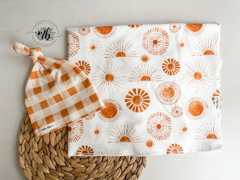 Puede incluir: Un gorro de beb&eacute; de cuadros blancos y naranjas con un nudo en la parte superior y una manta de arrullo blanca con un patr&oacute;n repetido de soles naranjas. El texto "maple bliss" est&aacute; en la etiqueta de la manta.