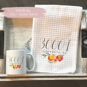 May include: A white mug and waffle-weave towel with the text "30001 ALPHARETTA, GA" and peach illustrations. The items are displayed on a distressed white wooden tray. The image also includes the text "YOUR zip, city + state here!"