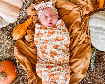 May include: A baby swaddled in a white swaddle with a pumpkin print, wearing a white headband with a bow. The baby is lying on a brown blanket surrounded by pumpkins and gourds.
