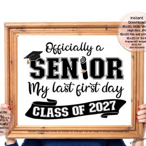 May include: A rectangular wooden frame containing a white sign with black text that reads Officially a SENIOR My last first day CLASS OF 2027. The design includes a black graduation cap icon and a rolled diploma icon replacing the letter I.