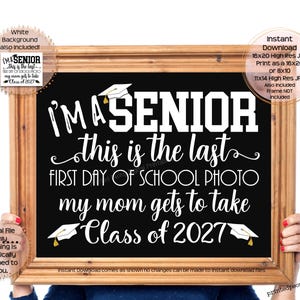 May include: A rectangular wooden frame holding a black sign with white text that reads I'M A SENIOR this is the last FIRST DAY OF SCHOOL PHOTO my mom gets to take Class of 2027, featuring white graduation cap illustrations at the top and bottom.