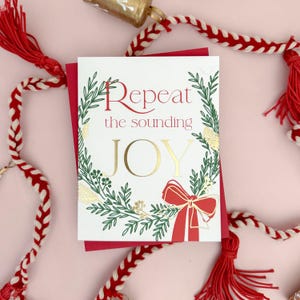May include: A Christmas greeting card with the words "Repeat the sounding JOY" in red and gold. The card features a green wreath design with a red bow. The card is on a red background with red and white rope and tassels.