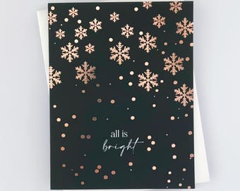 Può includere: Un biglietto nero con un design di fiocchi di neve e punti in foglia d'oro rosa. Il testo "all is bright" è scritto in bianco sul biglietto.