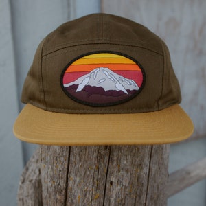 May include: A green and brown baseball cap with a mountain and sunset patch. The patch is a circle with a white mountain, a red, orange, and yellow sunset, and a brown base.
