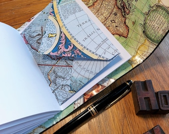 Op de afbeelding: Een vintage-stijl notitieboek met een wereldkaart omslag. Het notitieboek is open om lege pagina's te onthullen en een kaartdetail op de binnenkant van de omslag. Een zwarte pen en houten letterblokken zijn ook in de afbeelding te zien.