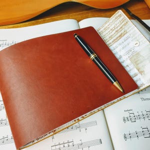 May include: A brown leather-bound journal with a gold pen lying on top of it. The journal is open to a page of sheet music with the word "Andante" printed on it.