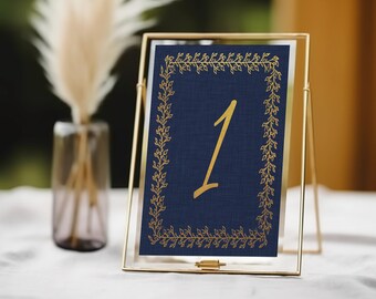 May include: Gold table number card with a navy blue background and a gold floral border. The number "1" is written in gold script.