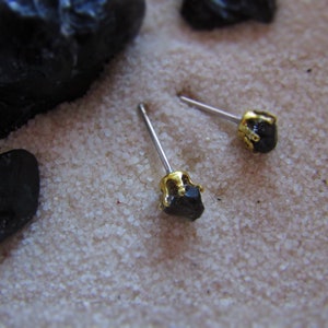 May include: A pair of black gemstone earrings with gold settings. The earrings are on a white sandy surface.